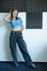 Teen girl in a blue crop top and cargo pants, wearing headphones in a school soundproof room. Concept of school media club, teen creativity, and student audio or podcast recording