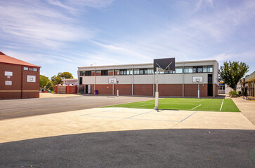 Outdoor basketball courts at a public primary school in Australia, with asphalt and synthetic turf surfaces, surrounded by buildings on campus. Educational infrastructure or elementary school facility