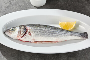 Whole sea bass on white oval plate, bright overhead, minimal stone background