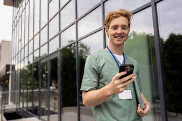 Caucasian medical worker in uniform standing by clinic wall using internet on smartphone