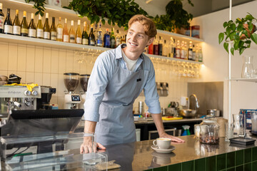 Smiling male coffee shop worker in apron standing at counter near espresso machine