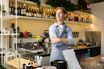 Cheerful man in apron posing near espresso bar standing with arms crossed in coffee shop interior