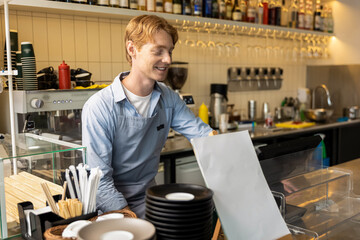 Pleased barista leaning slightly forward on bar counter smiling toothily standing inside coffee shop