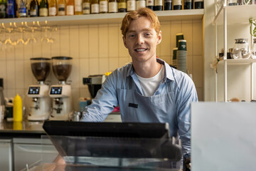 Joyful male café worker standing behind register wearing apron preparing order at espresso bar