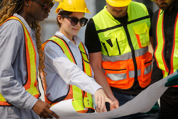 Diverse team of construction workers wearing safety helmets and reflective vests looking blueprint discussing building plans on construction site for teamwork and project planning concepts.