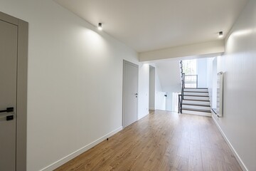 Bright, modern hallway with wood floors, white walls, and minimalist lighting. A staircase leads to an upper level, enhancing the spacious feel
