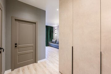 modern hallway featuring a closed taupe door, a partial view into a living room with a grey sofa and green curtains, and a beige closet with black handles