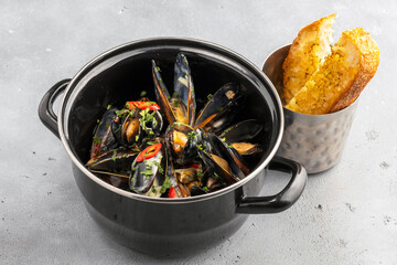 Black pot filled with cooked mussels in creamy sauce, garnished with fresh parsley and red chili slices, served alongside crispy toasted bread in a metal cup, on a light stone background.