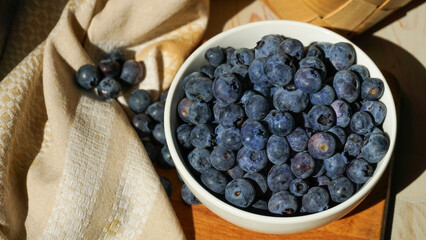 A bowl of ripe blueberries sits on a wooden board with a woven basket in the background, creating a rustic and natural setting ideal for showcasing healthy, fresh, and organic food. Close up