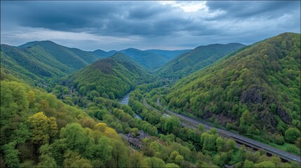 Fototapeta premium Verdant valley, winding road, and brooding sky. A high-angle view of a lush valley, with a river snaking through forested hills. Highway cuts through the green landscape. Dark clouds hover above