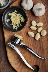 Top view of garlic grinder and plate with crushed garlic and fresh garlic cloves on wooden cutting board on wooden table.