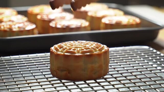 Traditional mooncake fresh from oven placed on cooling rack with tongs. Warm pastry with steam rising, more mooncakes in background.