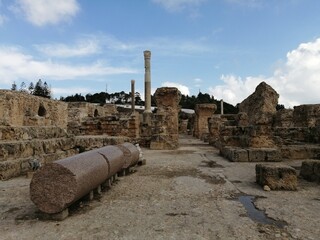 Ancient ruins with weathered stone columns and walls stand under a partly cloudy sky, preserving...