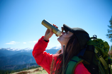 Woman in hiking gear taking a sip of water during mountain hike. Sporty female standing in front of snowy peaks.
