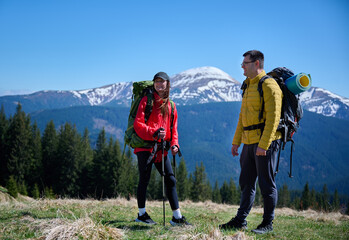 Two hikers taking a break on a mountain ridge. Sporty couple with hiking backpacks standing on background of snowy mountains.