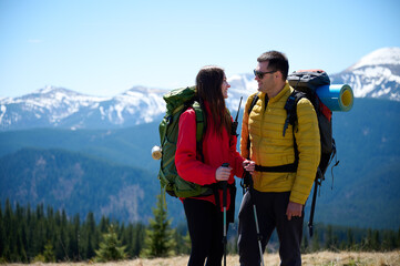 Two hikers taking a break on a mountain ridge. Sporty couple with hiking backpacks standing on background of snowy mountains.