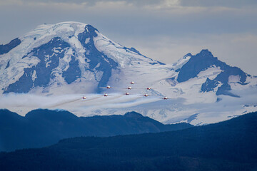 Nine aerobatic jets skim past a glaciated alpine peak, smoke streaming above forested hills, a...