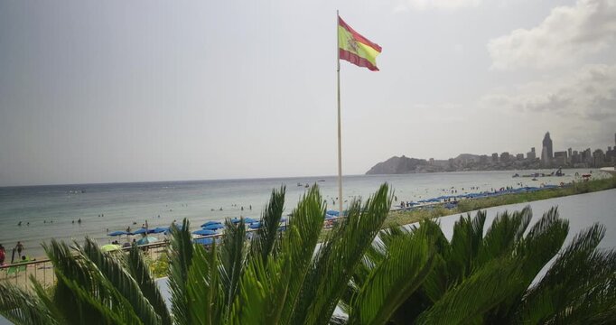 A Spanish flag waves above a crowded beach with blue umbrellas and swimmers, with the skyline of Benidorm visible in the distance. The scene captures summer energy and coastal beauty.