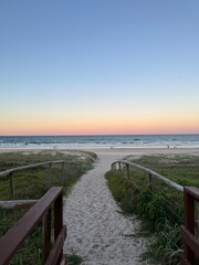 Obraz premium Wooden pathway leading through soft white sand dunes towards the open ocean at sunset, symbolizing peace, escape, and the beauty of untouched coastal landscapes.