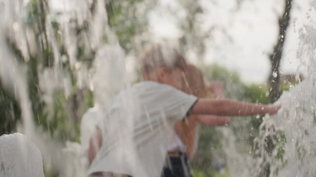 young playmates touching and playing with water from fountain in park, summer laughter and splashes, blurred background greenery, joyful energy and curiosity of children exploring cool droplets