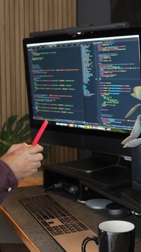 A programmer uses a smartphone with a large monitor displaying colorful code, highlighting a digital heart icon. Moody lighting and a modern setup create a tech-savvy ambiance. Vertical video format.