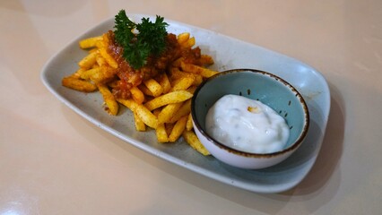 A serving of crispy fried potatoes garnished with herbs and sauce, accompanied by a creamy dip on a rectangular plate.
