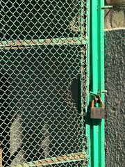 A close-up shot of a bright green chain-link fence with a rusted padlock, showing the texture of the weathered metal and the rough background behind it.