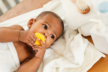 Close-up of an adorable baby wrapped in a towel after bath time, chewing on a yellow toy. Surrounded by soft towels and bath products, capturing a tender moment of care, comfort, and early childhood.