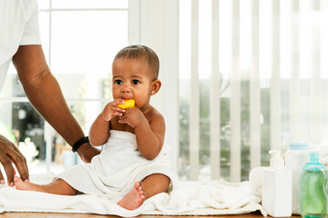 Adorable baby wrapped in a towel after bath time, chewing on a yellow toy, with parent nearby. Bright natural light and bath essentials create a warm, authentic family and childcare moment.