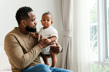 Loving father holds his baby girl near a bright window, sharing a tender moment. An authentic diversity family portrait symbolizing fatherhood, care, bonding, and Father’s Day celebration.