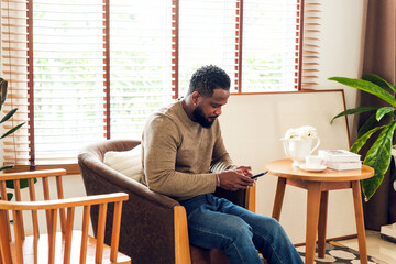 Relaxed man sitting in a cozy living room, focused on his smartphone. Bright natural light, indoor plants, coffee cup, and books create a warm, authentic lifestyle and home comfort atmosphere.