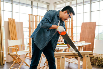 Focused carpenter in denim shirt and gloves cutting wood with a hand saw on a workbench, showcasing skilled craftsmanship, dedication, and authentic woodworking in a bright workshop.