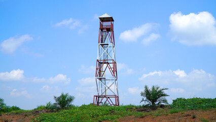 Metal observation tower standing tall against a bright blue sky, surrounded by green vegetation and palm trees, showcasing rural industrial infrastructure