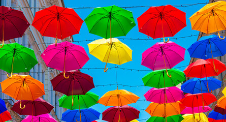 Multicolored open umbrellas above a beautiful street as decorations. Colorful umbrellas on the blue sky background.  Hanging rainbow umbrellas over clear blue sky. Abstract color background
