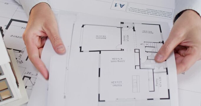 Man office worker in shirt flips through stack of apartment sketches and pauses at guest room. Experienced architect begins explaining position of furniture in lounge