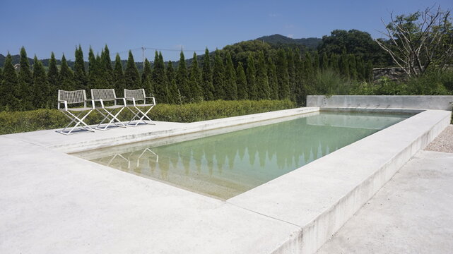 Simple outdoor swimming pool with three chairs on the side and trees in the background under blue sky