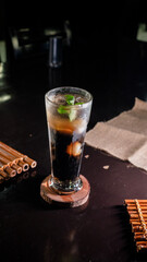 A tall glass filled with iced black coffee mixed with jelly cubes and topped with fresh mint leaves, placed on a round wooden coaster on a dark reflective table, creating a refreshing and sophisticate