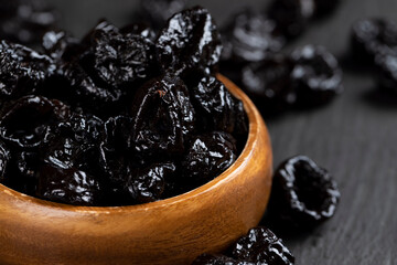 a wooden bowl filled with dried prunes, dark dried plum fruits poured into a bowl on black slate