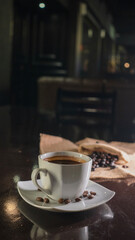 A white ceramic cup filled with rich black coffee, placed on a square white saucer with scattered coffee beans, set on a dark reflective surface with a blurred background of coffee beans on burlap.