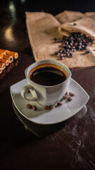 A white ceramic cup filled with rich black coffee, placed on a square white saucer with scattered coffee beans, set on a dark reflective surface with a blurred background of coffee beans on burlap.