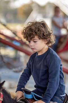 Curly-haired boy rides pony, holding saddle at fair