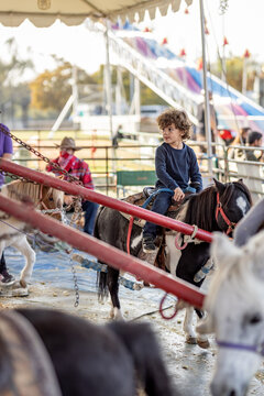 Curly-haired boy rides pony carousel under sunny tent