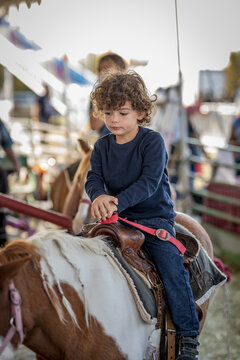 Young boy sits on brown and white pony at outdoor fair.
