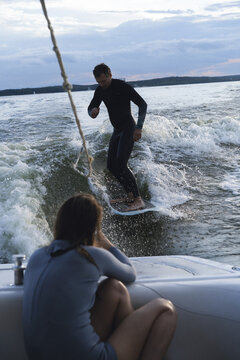 Man wake surfing on lake while woman films him during sunset