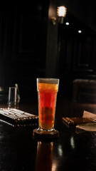 A tall glass of iced lychee tea with a vibrant orange gradient, containing whole lychee fruits at the base, served on a wooden coaster over a dark reflective surface, creating a refreshing and tropica