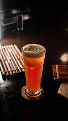 A tall glass of iced lychee tea with a vibrant orange gradient, containing whole lychee fruits at the base, served on a wooden coaster over a dark reflective surface, creating a refreshing and tropica