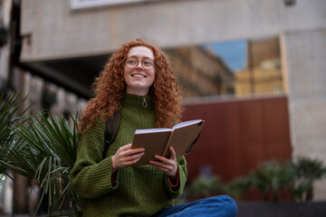 Red haired student with book and headphones sitting outdoors in city