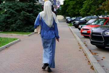 Woman in blue outfit walking along a city sidewalk with parked cars and greenery around in a...