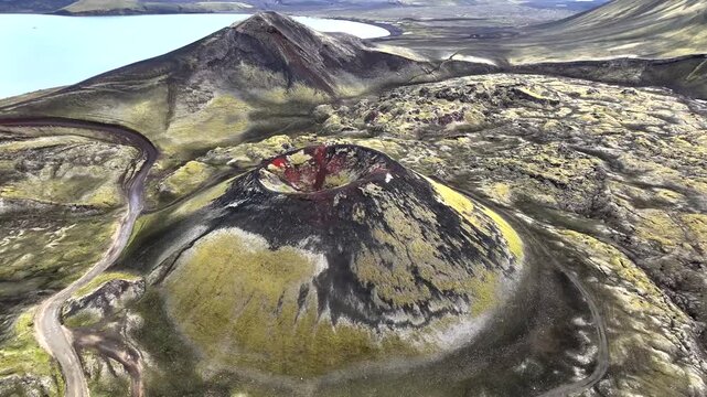 Aerial view of a volcanic crater with vibrant red interior and black rocky exterior, surrounded by green moss and a blue lake, Rang&aacute;r&thorn;ing ytra, Iceland.
