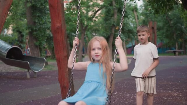 beautiful girl on wooden swing with chain handle smiling while playmate pushes from behind in green park setting, capturing joyful sibling interaction and motion blur in warm evening light playground